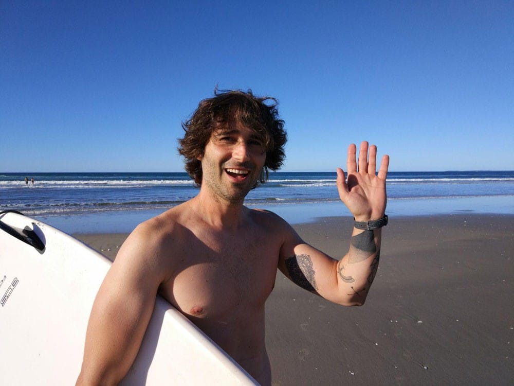 estudiante Nueva Zelanda chico moreno con el pelo rizado saludando a cámara y lleva una tabla de surf